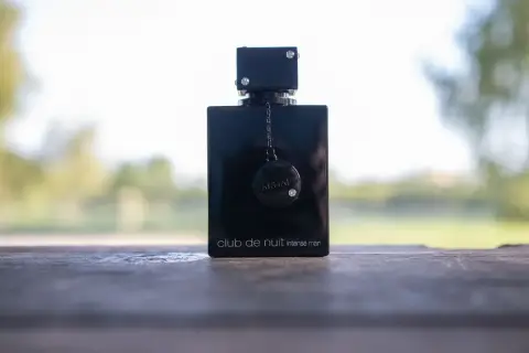 Elegancka czarna butelka perfum Club de Nuit Intense Man, ozdobiona łańcuszkiem i dyskiem z logo, stoi na drewnianym stole.